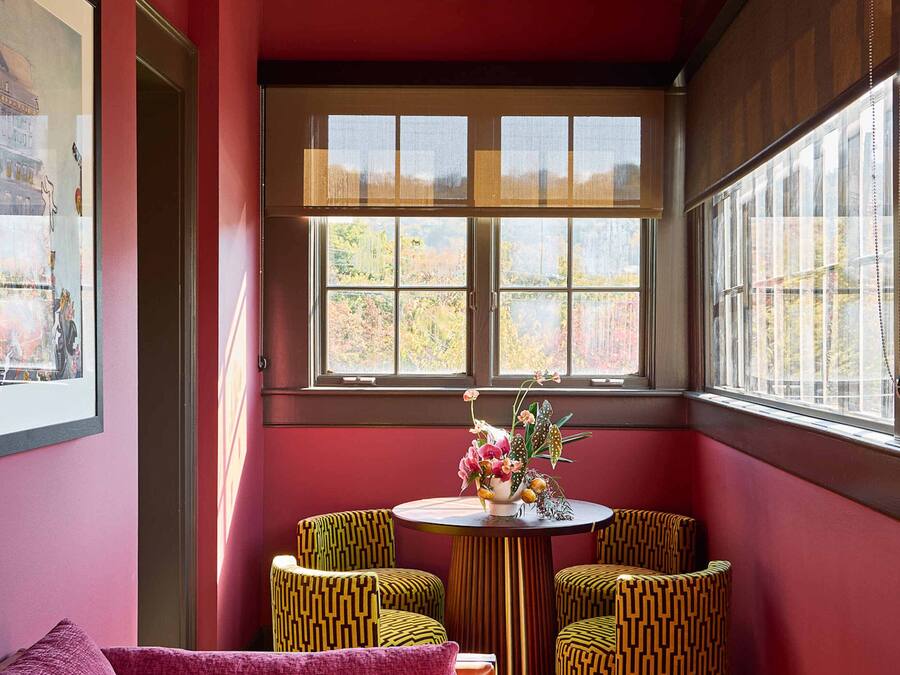 Eine gemütliche Ecke mit roter Wand, einem gemusterten Sofa, einem kleinen runden Tisch mit Stühlen und einem großen Fenster mit Blick auf Bäume.