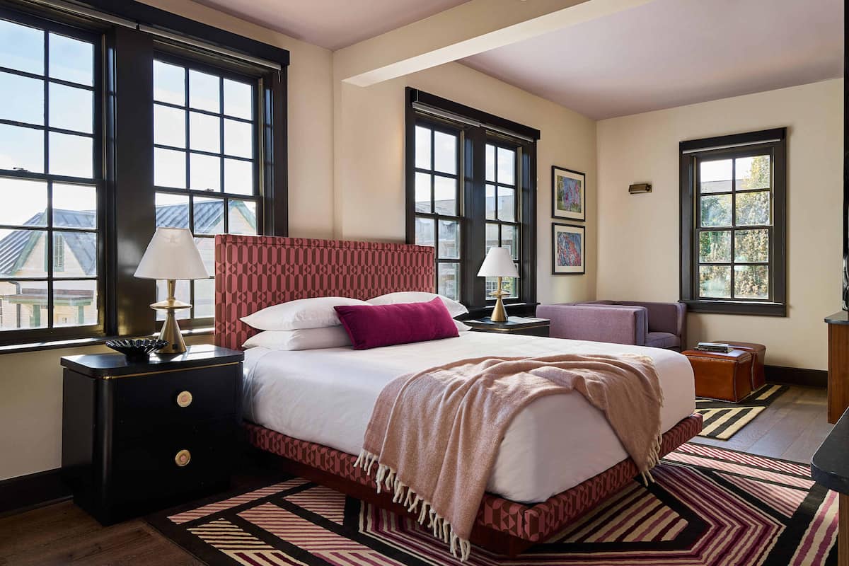 A bedroom with a large bed, two bedside tables, a nightstand, a chair, and a desk. There are large windows providing natural light and views of the outside.