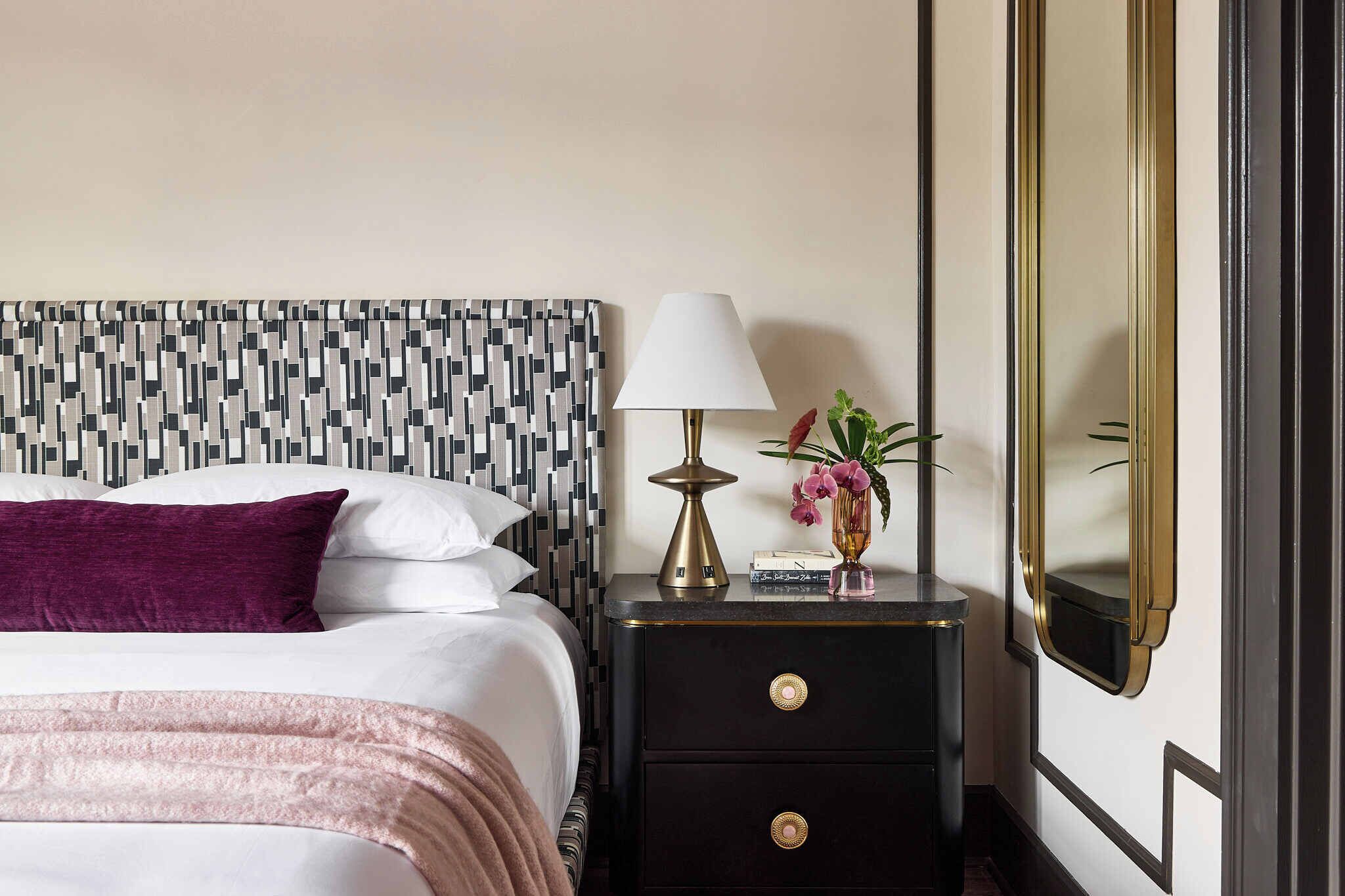 A neatly made bed with a patterned headboard, a bedside lamp, a small vase with flowers, and a mirror on a dark nightstand.