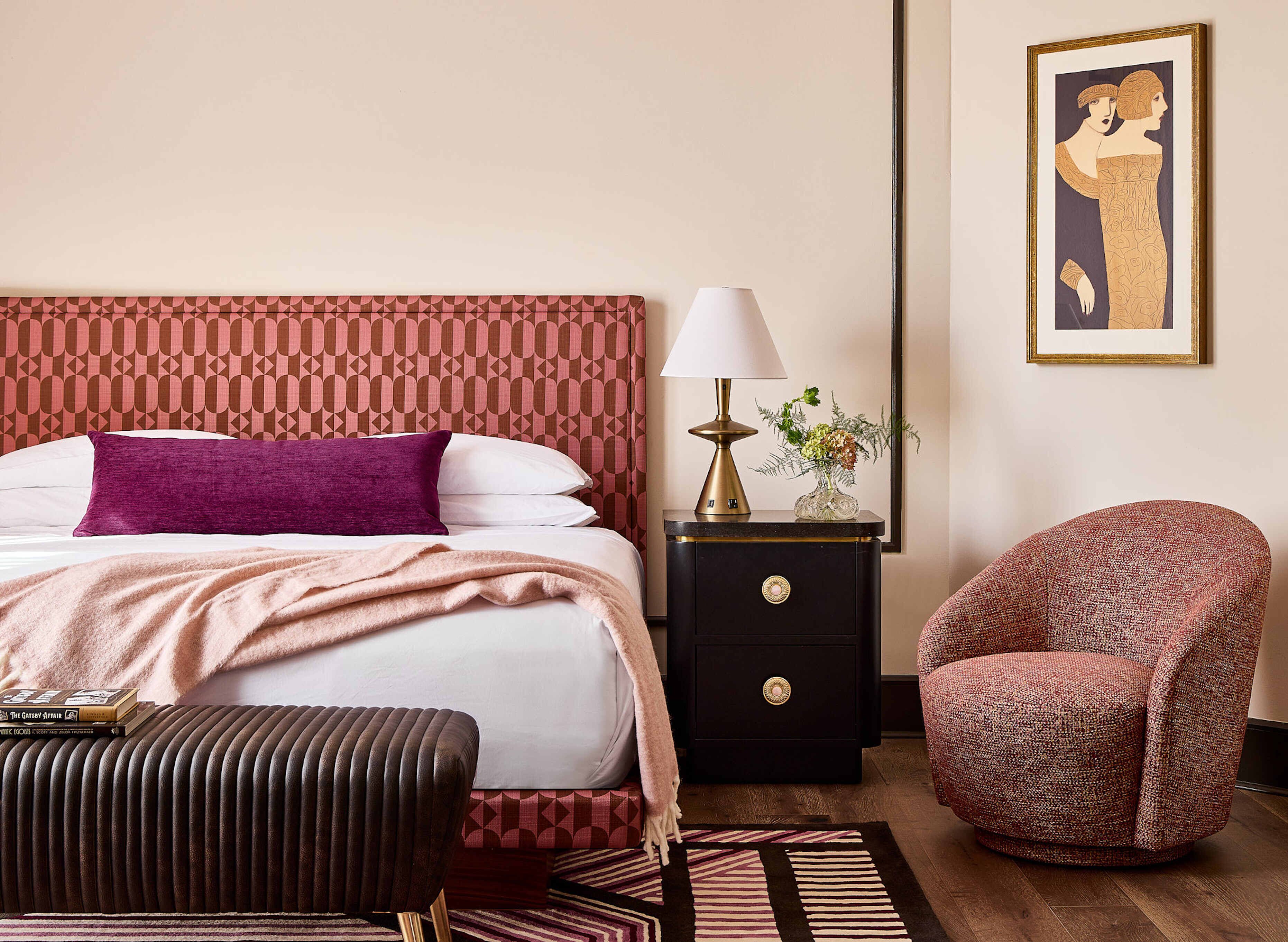 A hotel room with a patterned headboard, a bed with white linens, a purple pillow, a bedside table with a lamp and vase, a chair, and a framed artwork on the wall.
