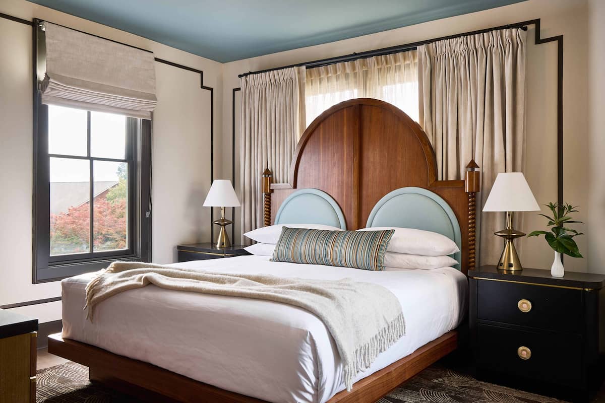 A bedroom with a large bed, two bedside tables with lamps, a wooden headboard, and a window with curtains.