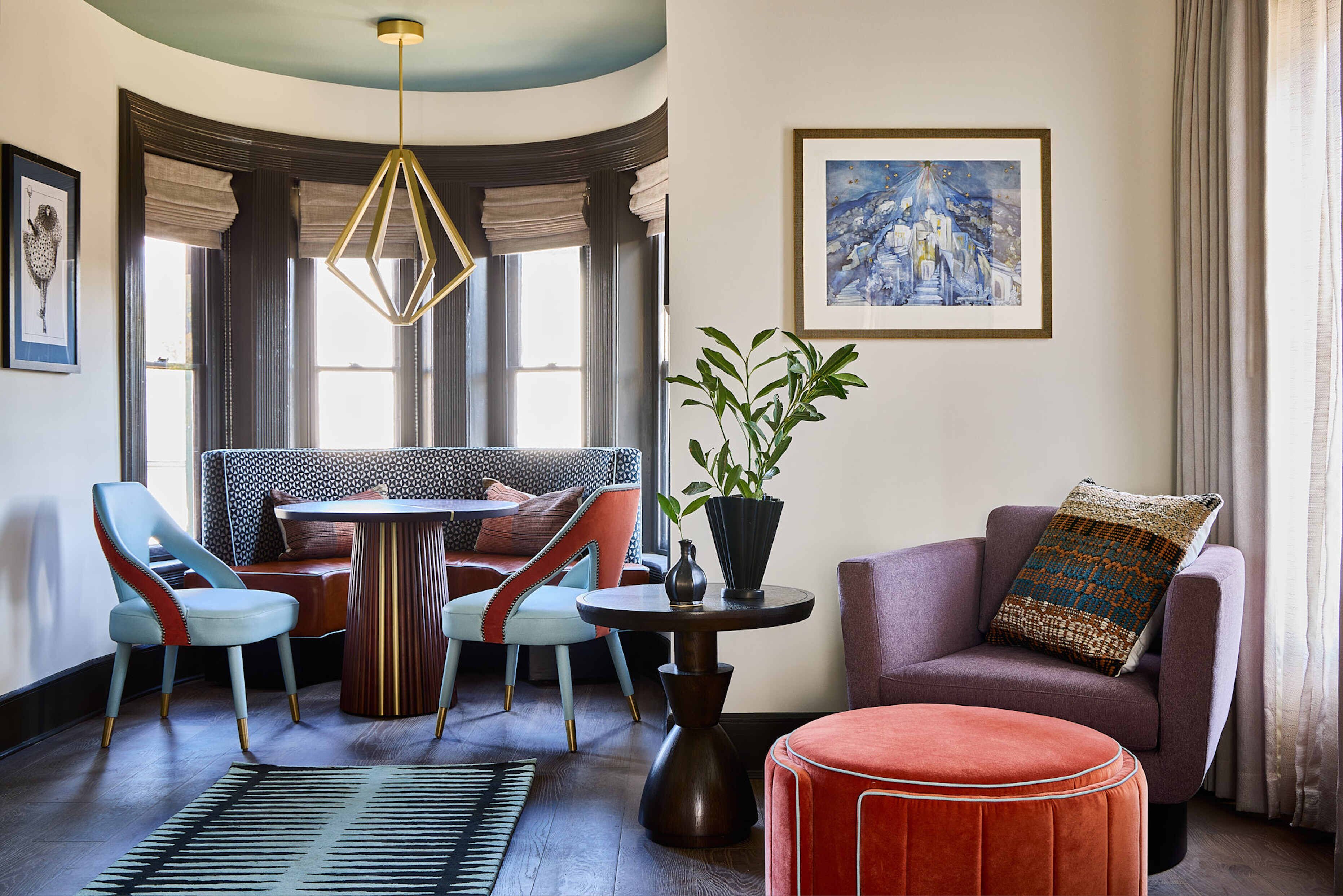 A modern living room with a round table, two chairs, a sofa, a red ottoman, a potted plant, and a framed painting on the wall.
