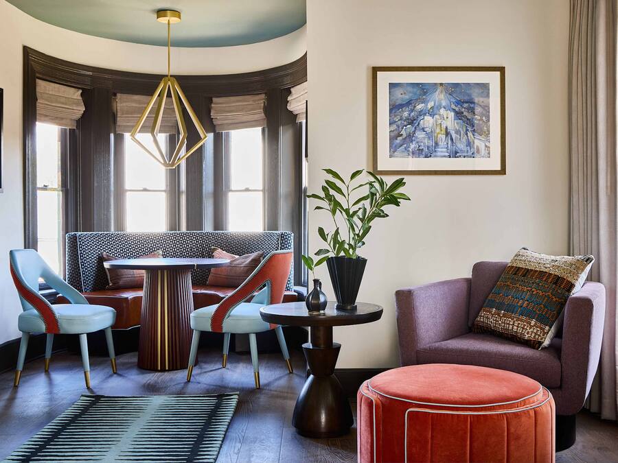 A modern living room with a round table, two chairs, a sofa, a red ottoman, a potted plant, and a framed painting on the wall.