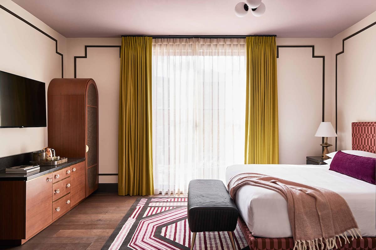 A hotel room with a bed, a TV, a wardrobe, a bedside table with a lamp, and a window with curtains.