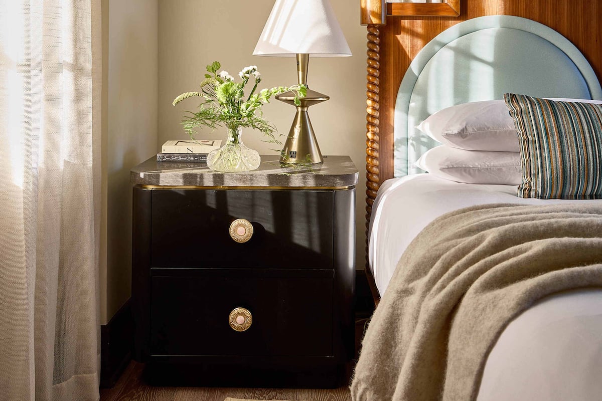 A neatly made bed with a striped pillow, a bedside table with a lamp and a vase of flowers, and a wooden headboard.