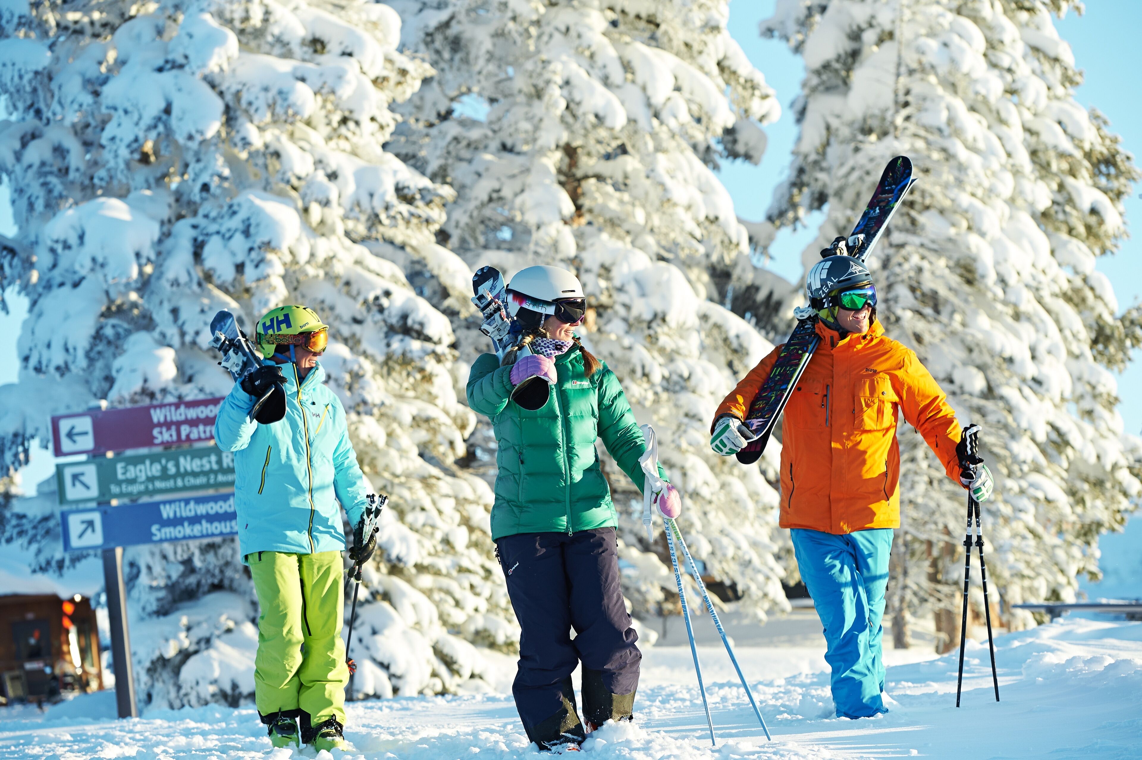 A snowy ski slope with skiers and a sign indicating directions to Wildwood Ski Patrol, Eagle's Nest, and Smokehouse.