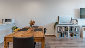 Un coin repas avec une table en bois, des chaises, un vase de fleurs, un four à micro-ondes posé sur une étagère et une bibliothèque remplie de livres.