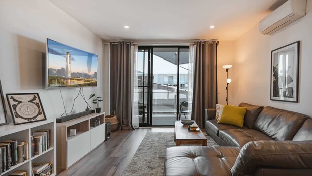 Un salon moderne avec un canapé en cuir, un téléviseur à écran plat fixé au mur, une bibliothèque et une vue sur un pont, visible à travers une porte vitrée.