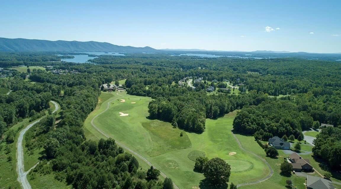 An aerial view of a golf course with a lake, forested areas, and residential properties.