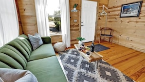 A cozy wooden cabin interior with a green sofa, a wooden coffee table with fruits and a bottle, and a view of the outdoors through a window.