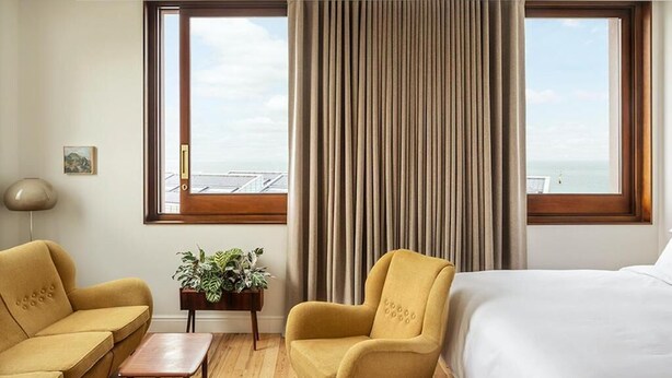 A hotel room with a bed, two armchairs, a small table, and a potted plant. There are two large windows with curtains, and a view of the sea.
