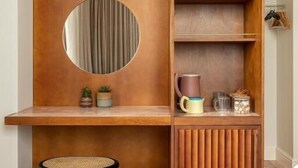 A compact room with a wooden desk, a round mirror, a stool, and a built-in shelf with various items.