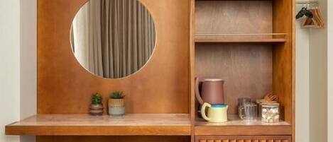A compact room with a wooden desk, a round mirror, a stool, and a built-in shelf with various items.