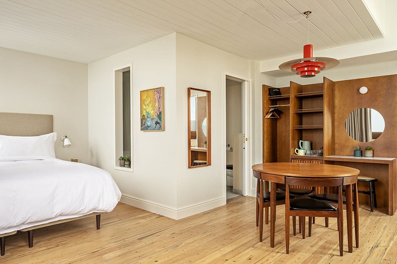 A hotel room with a bed, a dining table with chairs, a mirror, a wardrobe, and a red pendant light.