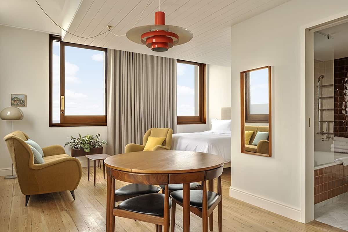 Habitación de hotel moderna con una cama grande, una mesa redonda de madera, dos sillones, una lámpara de pie y un ventanal con vistas a la ciudad.