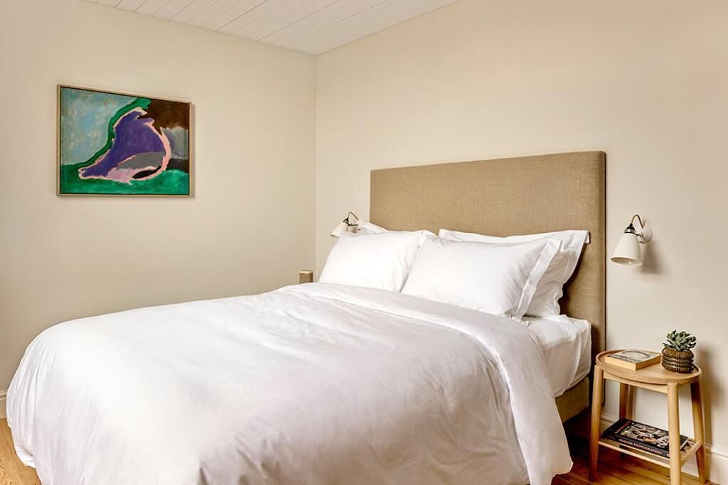 A neatly made bed with white linens in a room with a beige headboard. There is a small wooden side table with a lamp and a book, and a painting on the wall.