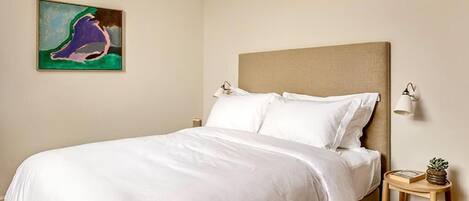 A neatly made bed with white linens in a room with a beige headboard. There is a small wooden side table with a lamp and a book, and a painting on the wall.