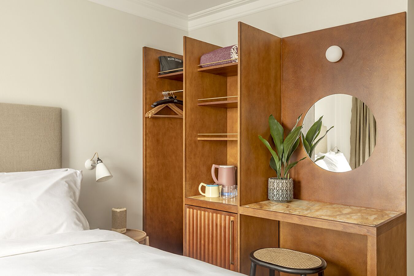 Quarto de hotel com cama, guarda-roupa com espaço para pendurar, espelho, mesa pequena com planta e uma cadeira.