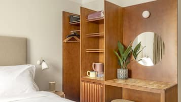 Quarto de hotel com cama, guarda-roupa com espaço para pendurar, espelho, mesa pequena com planta e uma cadeira.