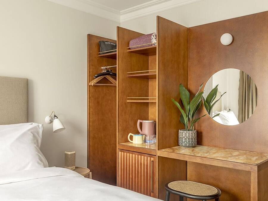 Quarto de hotel com cama, guarda-roupa com espaço para pendurar, espelho, mesa pequena com planta e uma cadeira.
