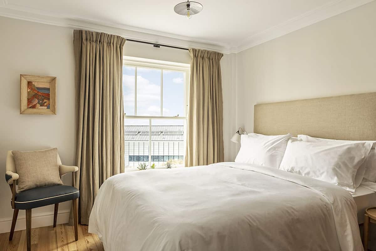 Habitación de hotel con una cama grande, una silla, una mesita de noche, una lámpara y un ventanal con cortinas.