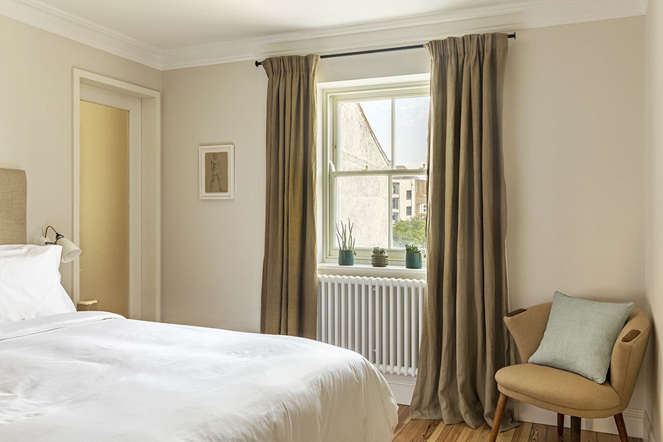 A hotel room with a bed, a chair, a window with curtains, a radiator, and a framed picture on the wall.