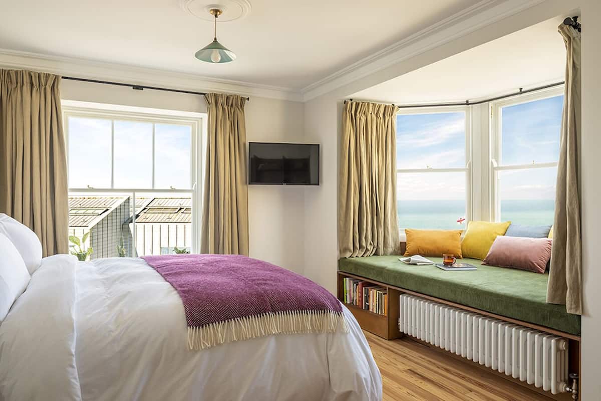 Un dormitorio con cama, un sofá cama verde con almohadas, una estantería, un televisor montado en la pared y vistas al mar a través de un ventanal.