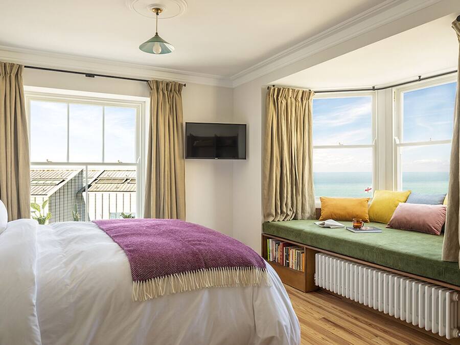 A bedroom with a bed, a green sofa bed with pillows, a bookshelf, a TV mounted on the wall, and a view of the sea through a bay window.