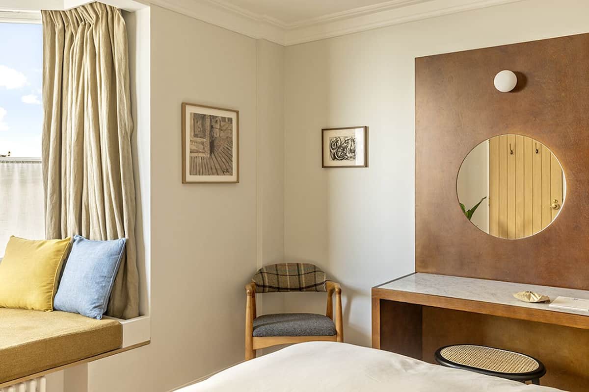 Habitación de hotel con cama, una silla, escritorio y un espejo grande. Hay dos cuadros en la pared.