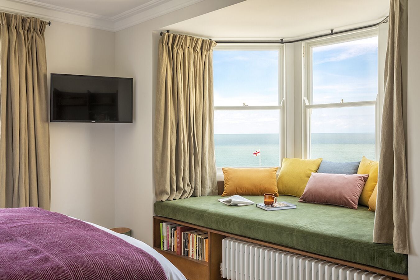 A room with a bay window overlooking the sea, a bed with a green bedspread, a bookshelf, and a flat-screen TV mounted on the wall.