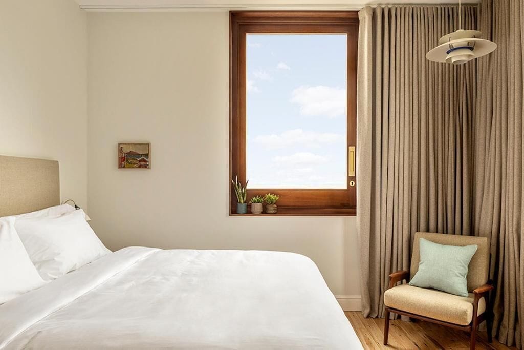 Ett hotellrum med en stor säng, en stol, ett fönster med gardiner och en liten hylla med växter.