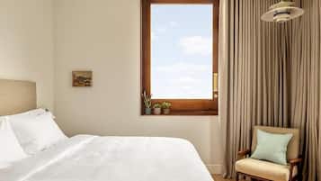 Ett hotellrum med en stor säng, en stol, ett fönster med gardiner och en liten hylla med växter.