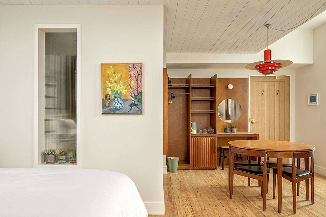 A modern bedroom with a bed, a wooden desk, a dining table with chairs, a closet, and a painting on the wall.