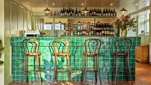 A bar area with a green tiled backsplash, wooden bar stools, and a variety of bottles on shelves.
