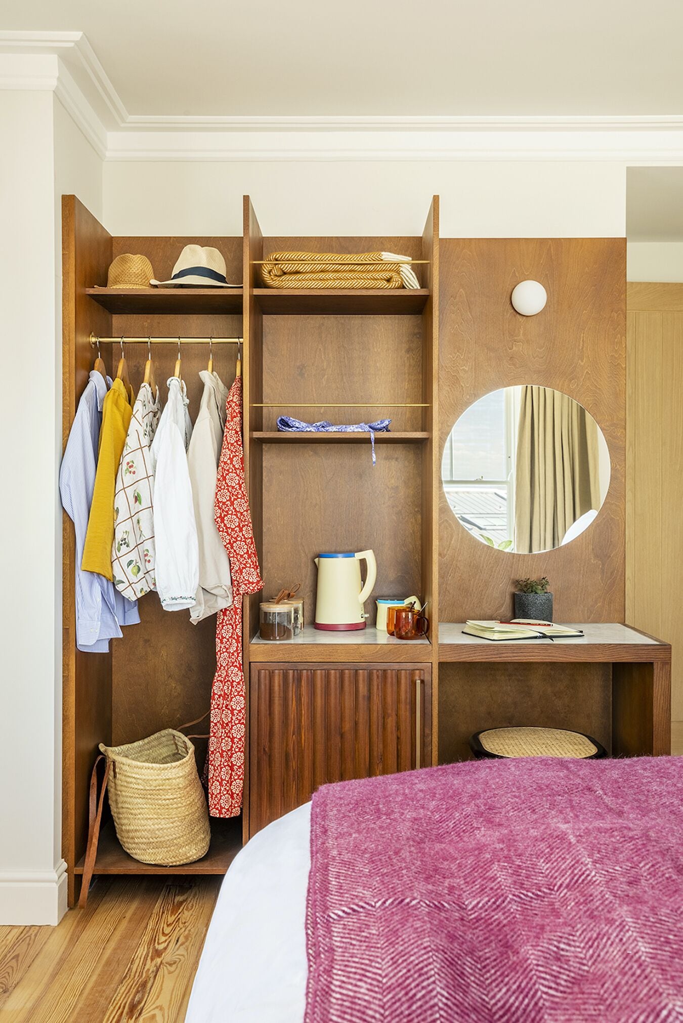 A bedroom with a wooden wardrobe, a bed with a pink blanket, a round mirror, and a small desk with a cup and a plant.