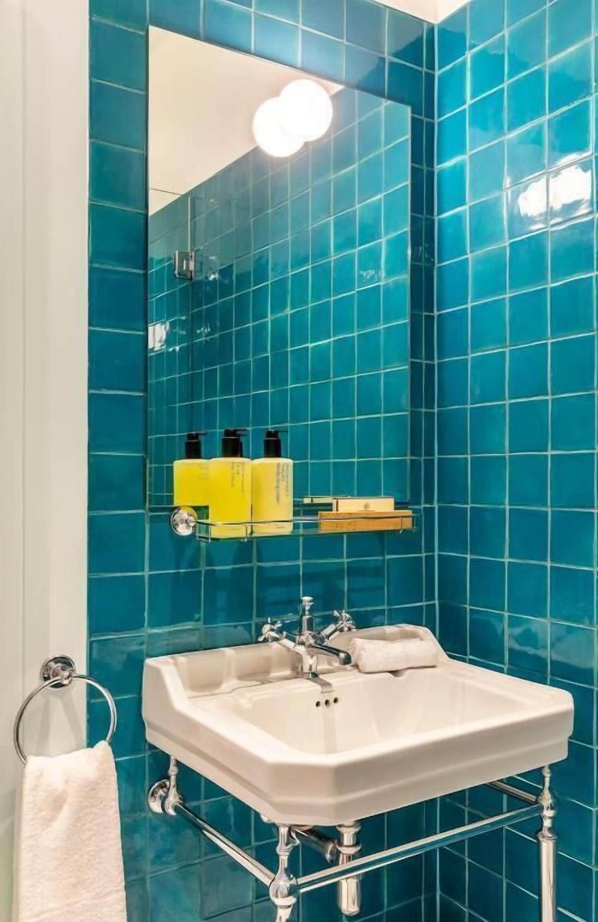 Un baño con lavamanos blanco, grifería cromada y un espejo encima. Los azulejos azules cubren las paredes.