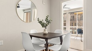 Un coin repas agrémenté d’un miroir rond, d’un vase avec des plantes et de chaises blanches disposées autour d’une table en bois foncé.