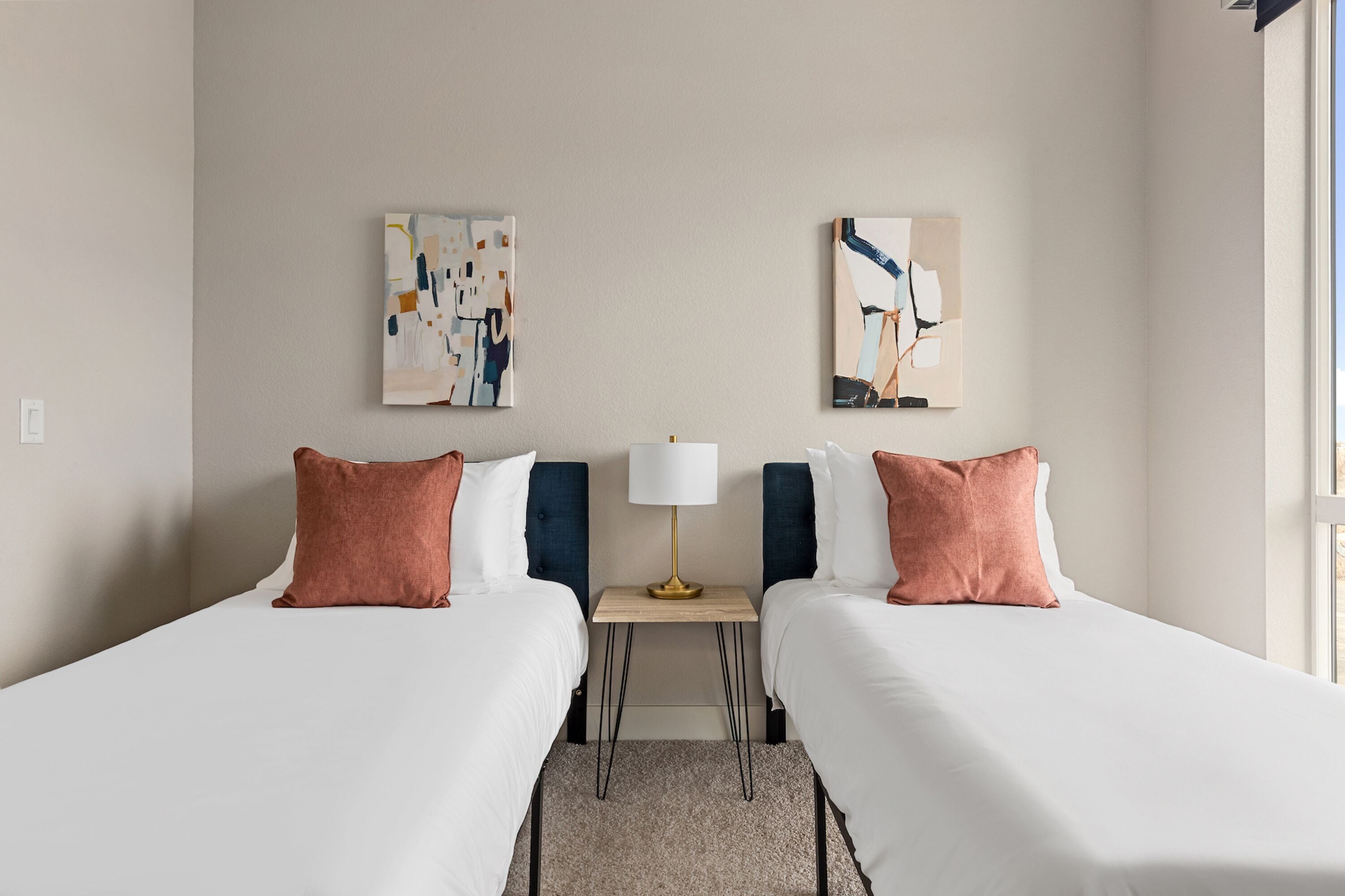 A hotel room with two beds, a small table with a lamp, and abstract artwork on the walls.
