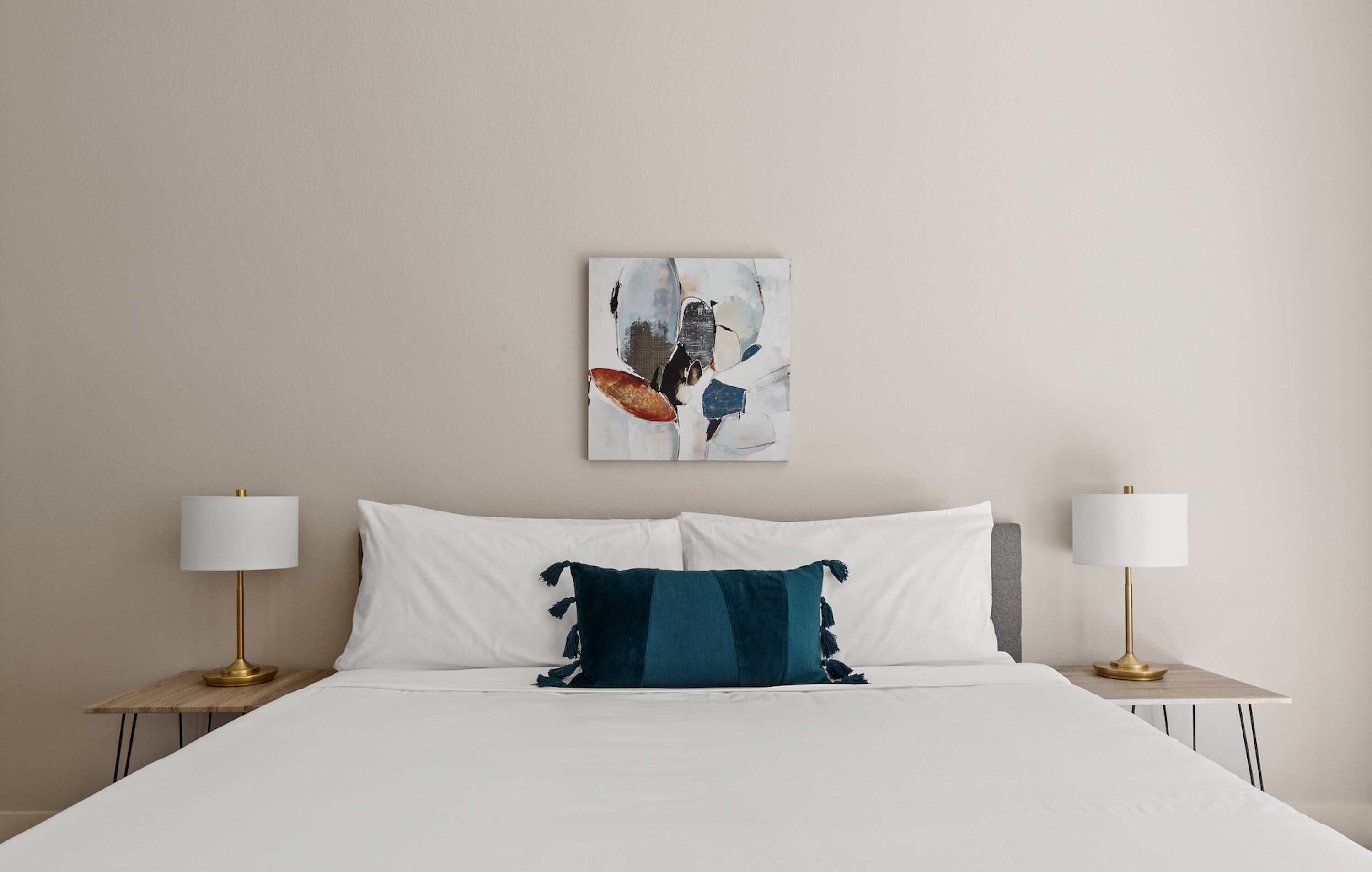 Un lit bien fait, agrémenté d’oreillers blancs et turquoise, deux tables de chevet avec des lampes, et un tableau abstrait accroché au mur.