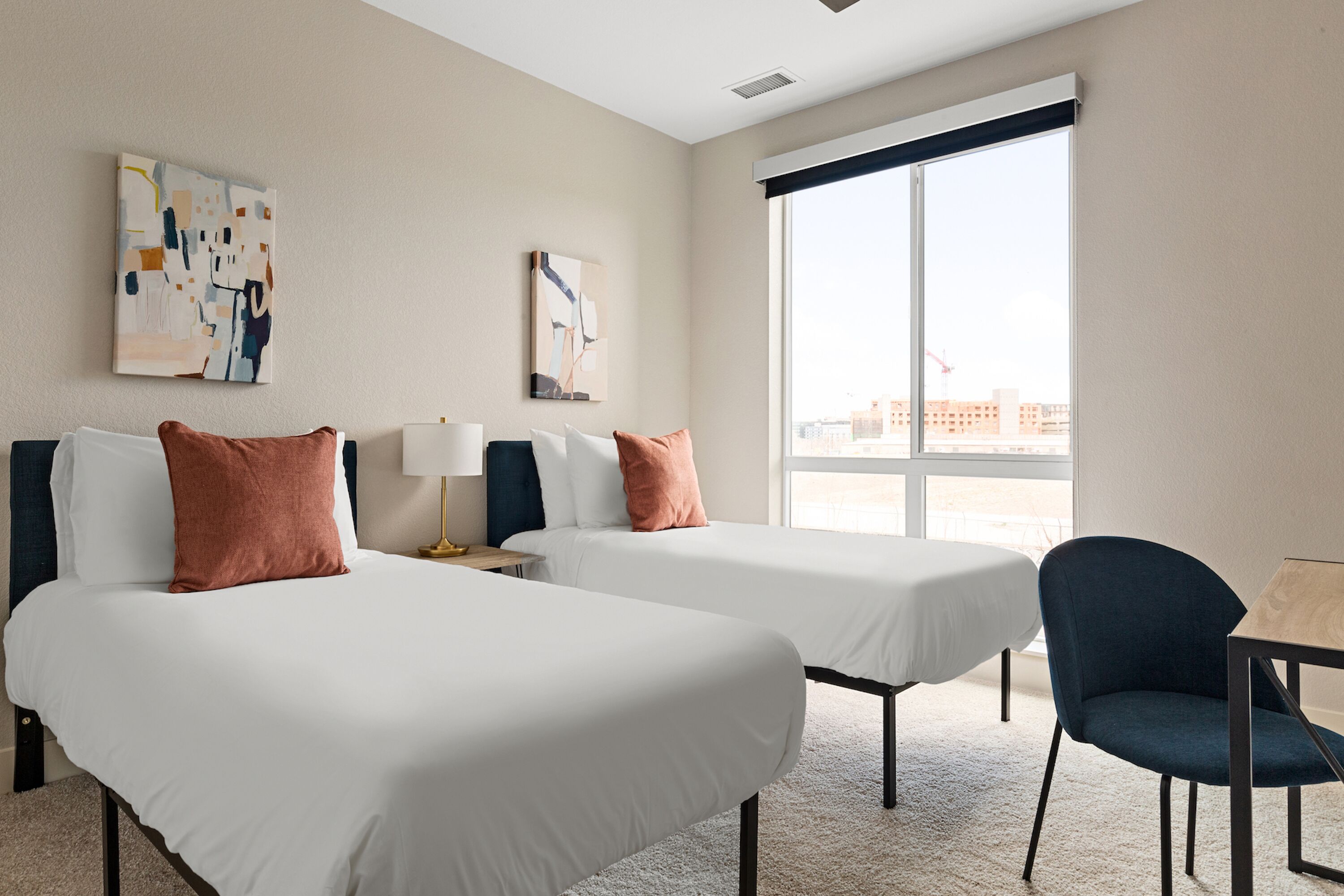 A hotel room with two beds, a desk, and a chair. There are two paintings on the wall and a large window.