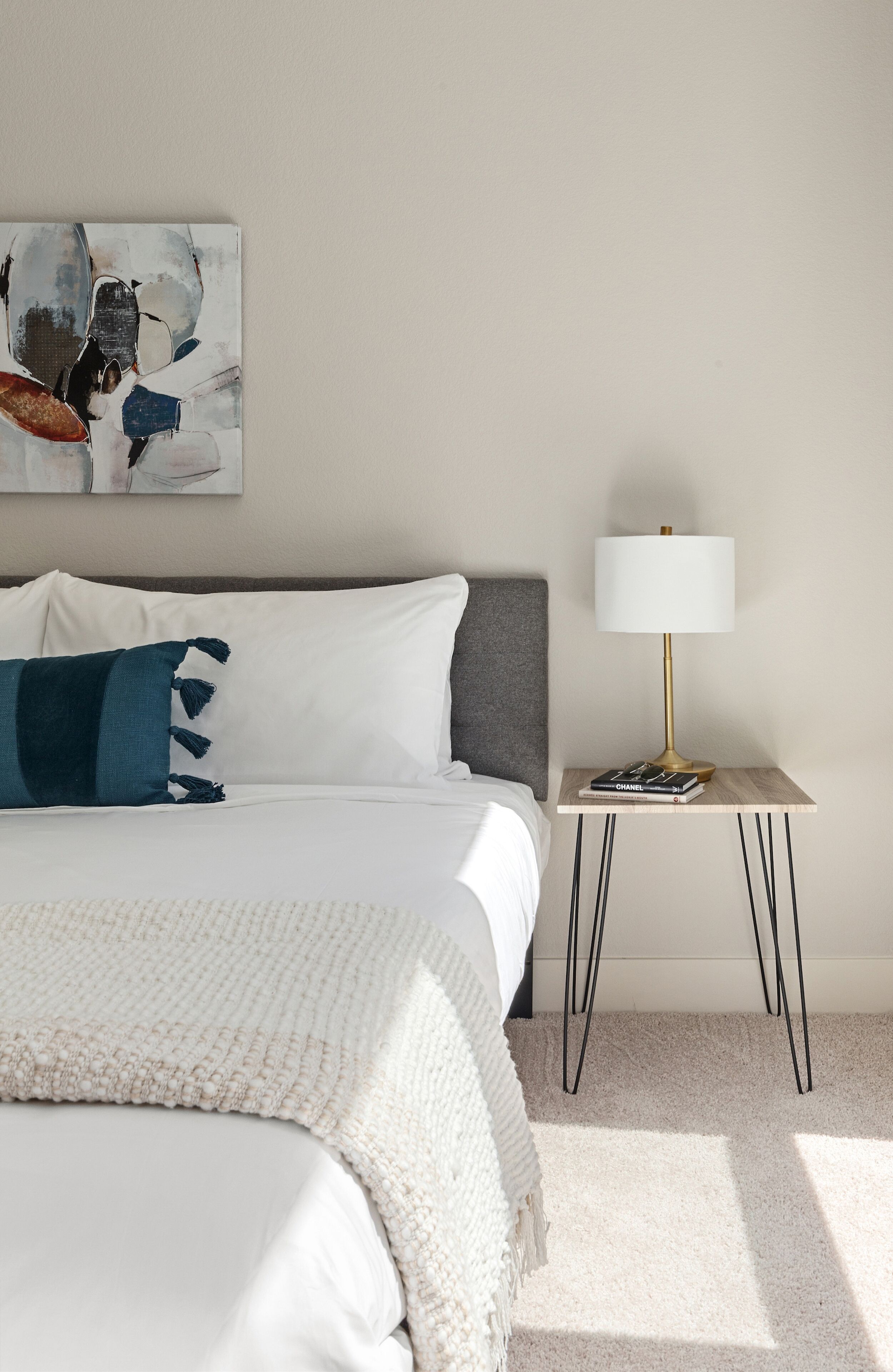 A neatly made bed with a white comforter and a blue throw pillow, a bedside lamp on a wooden nightstand, and a painting on the wall.