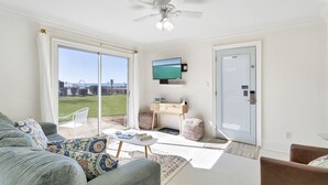 A living room with a sofa, coffee table, and a TV on a stand. There is a sliding glass door leading to a balcony with a view of the city. - Sea Cabins 2B - Newly Renovated, Heated Community Pool, Deeded Beach Access! (Miramar Beach)