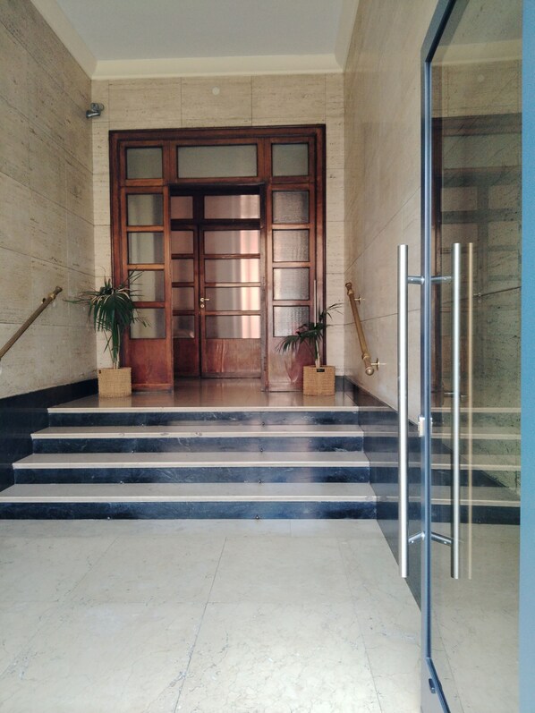 A marble-floored entrance with a wooden door and glass panels, flanked by potted plants and a metal railing.