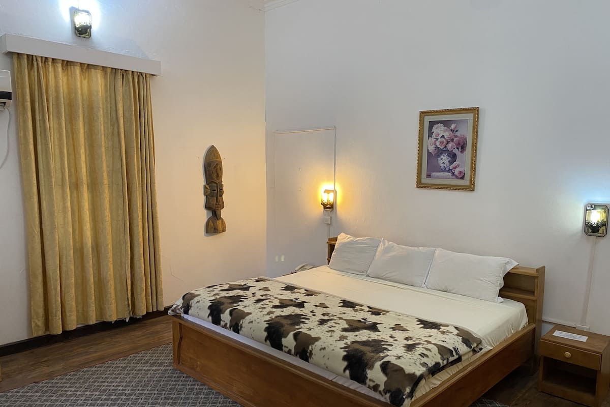Un dormitorio con cama de madera, una alfombra estampada, una lámpara de pared, un cuadro enmarcado y una cortina.