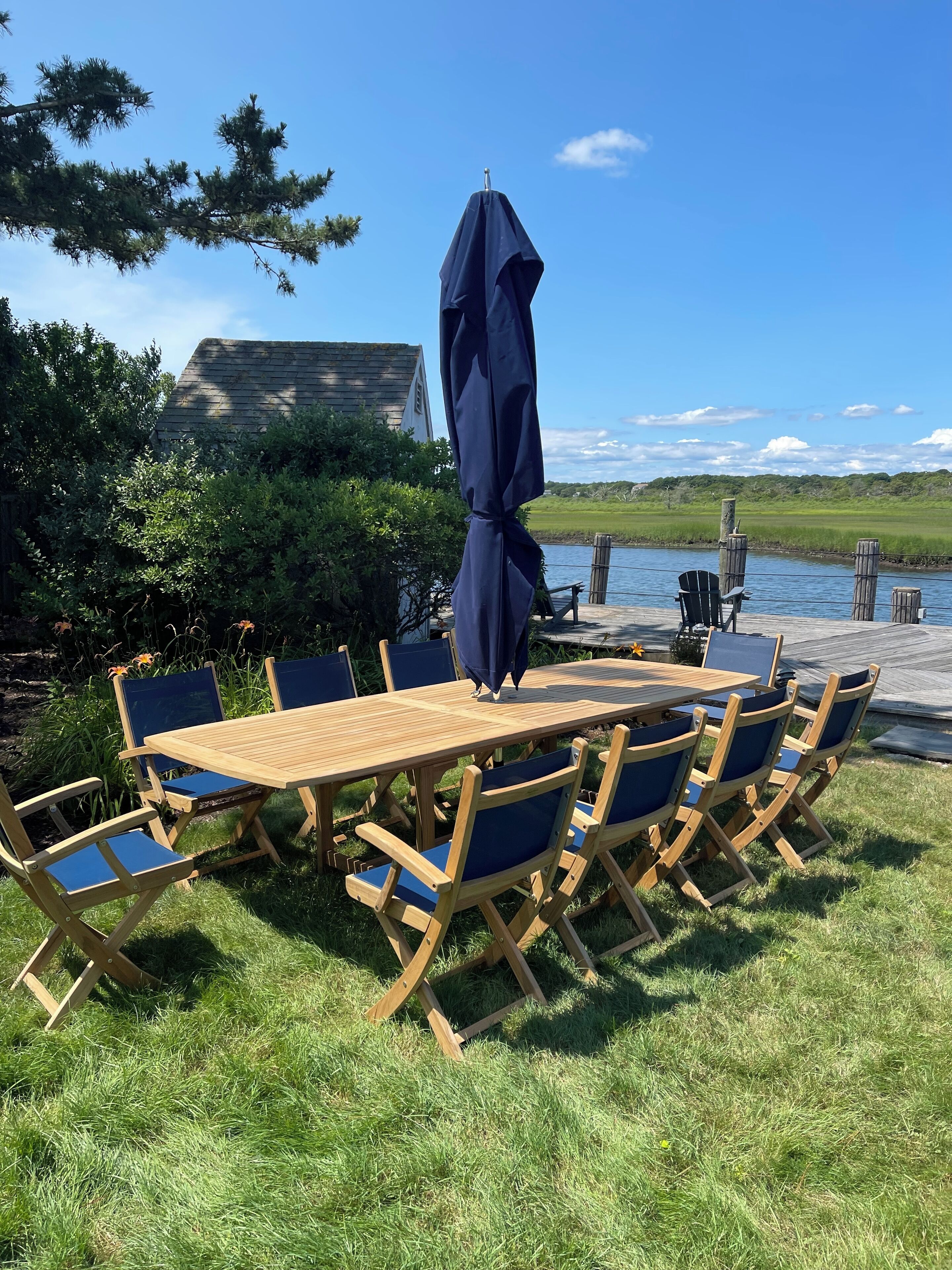 An outdoor dining area with a wooden table and blue chairs, a dark blue umbrella, and a view of a body of water and greenery.