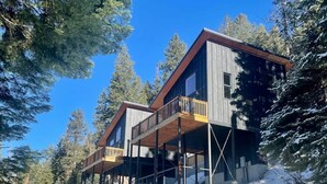 Une cabane moderne à deux étages, dotée d’une terrasse en bois et d’un balcon, entourée de neige et d’arbres.