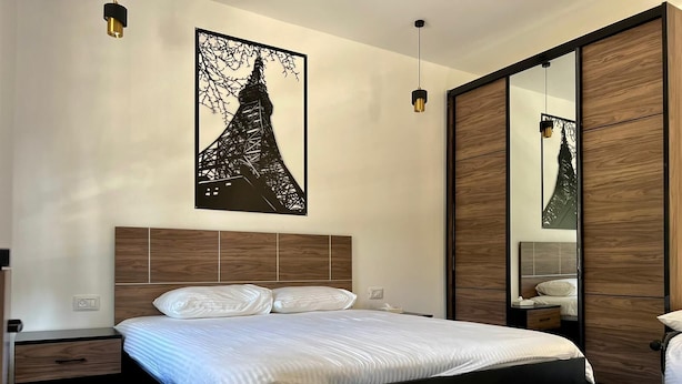 Quarto moderno com cama grande, cabeceira de madeira, espelho e um objeto decorativo na parede.