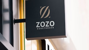 Uma placa preta com o logotipo e o nome "ZOZO GUESTHOUSE" fixada num edifício.