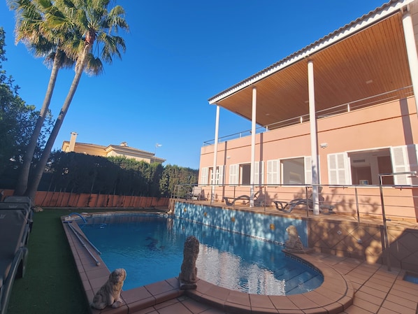 Une piscine ornée d’une statue de lion en pierre, un patio couvert et, au fond, un bâtiment avec un toit en tuiles.