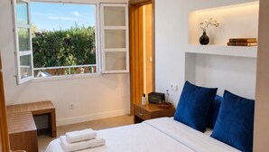 A bedroom with a large bed, bedside tables, a nightstand, a shelf with books and a vase, and a window with a view of greenery. - Villa Berria, 9 Pool / BBQ / Your Oasis near Palma (Es Pont d`Inca)